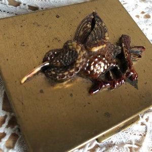 May include: A vintage brass compact with a detailed, three-dimensional hummingbird design. The bird is brown and tan with a long, light-colored beak, perched on a branch. The compact has a rectangular shape and a slightly worn, antique appearance.