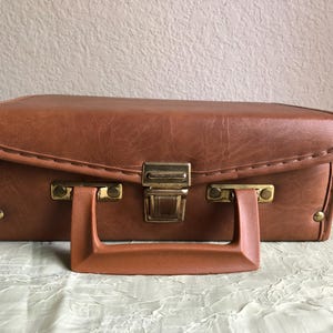 May include: A rectangular brown faux leather hard-shell case with a textured surface. The case features a sturdy matching brown plastic handle, gold-toned metal hardware, and a central latch mechanism for secure closure.