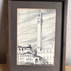 May include: A framed black and white architectural sketch. The artwork depicts a tall tower and buildings, with a cloudy sky background. The frame is dark brown.