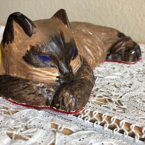May include: A ceramic figurine of a brown and white cat with blue eyes, lying down with its head resting on its paws. The cat is painted with realistic details and has a yellow and red base.