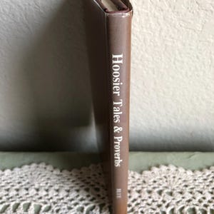 May include: A brown book titled "Hoosier Tales & Proverbs" with white lettering. The book's spine is visible, showing the title and author. The book is standing upright on a lace doily.