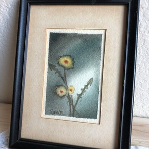 May include: A framed watercolor painting of three yellow dandelions with green stems against a gray background. The painting is signed "JIANNE" in the bottom left corner.