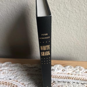 May include: A hardcover book titled "WHITE SHARK" by Peter Benchley. The book has a black cover with gold lettering and a series of gold dots. The book is standing upright on a white lace surface.