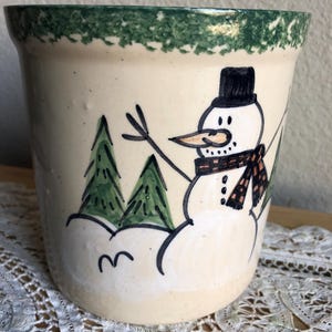 May include: Cream-colored ceramic pot with a hand-painted snowman design. The snowman wears a black top hat and a brown and black scarf. Green trees and a green rim complete the winter scene.
