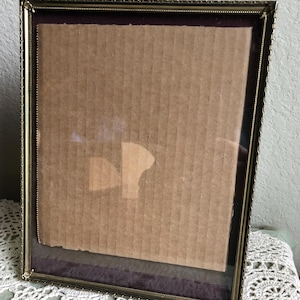 May include: A rectangular picture frame with a thin, metallic gold-toned border featuring a beaded edge. The frame contains a piece of light brown, vertically ribbed cardboard backing set against a dark purple velvet matting behind clear glass.
