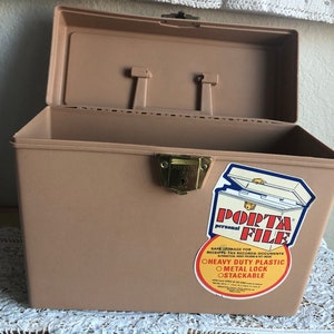 May include: Tan Porta File personal storage box with an open lid. The box has a metal lock and a sticker that reads "Porta File". The box is made of heavy-duty plastic and is stackable.
