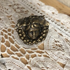 May include: A gold-toned metal decorative knob with a detailed design of a woman's face surrounded by grapes and leaves.