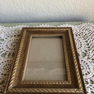 May include: A rectangular picture frame with a brown finish and gold-colored rope-like detailing. The frame has a simple, classic design, suitable for displaying photographs or artwork. The frame is sitting on a white lace doily.