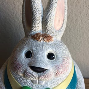 May include: A ceramic Easter bunny figurine with a white body, pink-lined ears, and a brown mustache. The bunny wears a teal vest with a yellow collar and a green bow tie. The cheeks are blushed pink, and the eyes are black.
