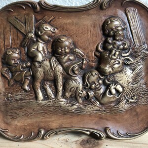 May include: A vintage wall plaque with a brown wooden frame and a raised relief design of children playing with animals. The plaque features a brown cow, a brown donkey, and five children in a rustic setting.