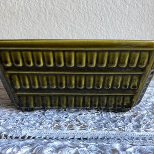 May include: A green ceramic planter with a geometric design. The planter has a rectangular shape with a slightly flared rim and a textured surface.