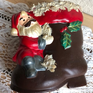 May include: A ceramic Santa Claus figurine holding a white object, sitting on a brown boot with green holly and red berries. The boot is decorated with white paint.