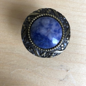 May include: A round, ornate, brass-colored knob with a blue and white stone inlay. The knob has a detailed, intricate design.