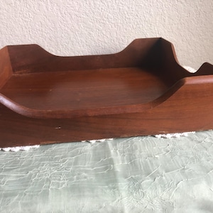 May include: A rectangular wooden desk tray with a medium brown finish. The tray features raised sides with a curved cutout design on the front and back edges, and visible finger joint construction details on the corners of the wooden structure.