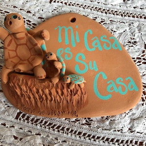 May include: A terracotta-colored plaque with three-dimensional turtle figures. The plaque has the Spanish phrase "Mi Casa es Su Casa" in turquoise script. The turtles are brown with blue accents. The plaque is on a white lace surface.