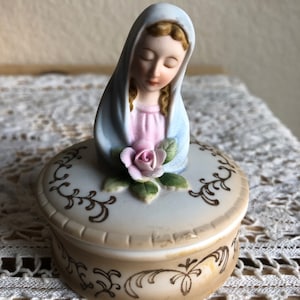 May include: A porcelain trinket box with a lid featuring a figurine of the Virgin Mary holding a pink rose. The box is decorated with a brown floral design.