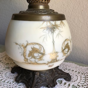 May include: Antique oil lamp with a white glass globe featuring gold bamboo and floral designs. The lamp has a bronze-colored metal base and top, showcasing intricate detailing. A vintage decorative piece.