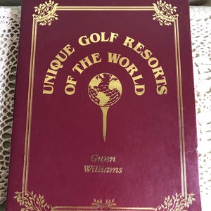 May include: A maroon book titled "Unique Golf Resorts of the World" with gold lettering and accents. The cover features a golf ball on a tee graphic. The author's name, Gwen Williams, is also displayed.