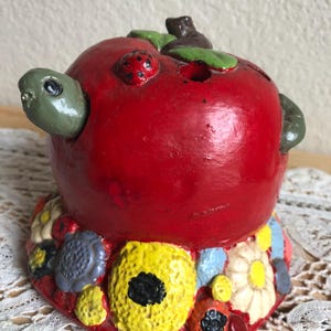 May include: A whimsical, hand-painted ceramic apple-shaped container. The bright red apple features a turtle head, a ladybug, and green leaves. The base is adorned with colorful, textured flowers and shapes, creating a unique decorative piece.