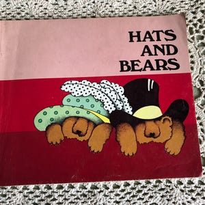 May include: A children's book titled "Hats and Bears" with a pink and red cover. The illustration shows two brown bears wearing hats, one a top hat and the other a polka-dotted hat. The book is on a white lace surface.