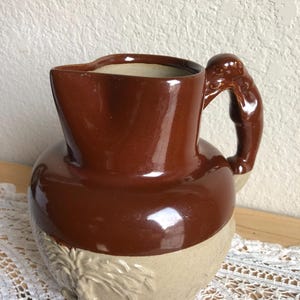 May include: A brown and beige ceramic pitcher with a sculpted handle. The pitcher has a brown glaze on the top half and a beige glaze on the bottom half. The bottom half has a sculpted design of a plant.