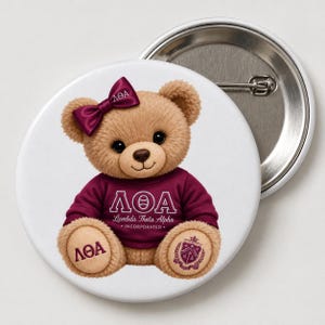 May include: A round pinback button featuring a tan teddy bear wearing a maroon sweater with the Greek letters ΛΘΑ and the text Lambda Theta Alpha Incorporated. The bear has a maroon bow with ΛΘΑ on its head, and its paws feature the letters ΛΘΑ and a crest.