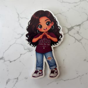 May include: A die-cut sticker featuring a cartoon character with long wavy dark hair, wearing silver hoop earrings, a maroon t-shirt with the text Lambda Theta Alpha 1975, blue distressed jeans with a knee rip, and white and maroon sneakers.