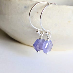 May include: A pair of silver hoop earrings with faceted, light purple gemstone drops. The earrings have a simple, elegant design, with the gemstones adding a touch of color and sparkle. The earrings are set against a soft, neutral background.