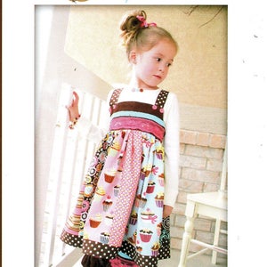 May include: A girl wearing a brown and pink cupcake-themed dress with a white long-sleeved shirt. The dress has a brown and white polka dot ruffle at the bottom. The dress is from Pink Fig Patterns and is called "The Cuppy Cake Dress" No. 11.