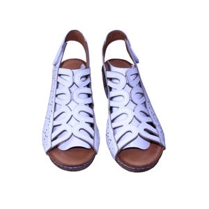 Puede incluir: Un par de sandalias de cuero blanco con un diseño abierto. Las sandalias tienen una correa trasera y una suela marrón. La parte superior presenta un patrón de corte decorativo. Los zapatos están sobre un fondo blanco.