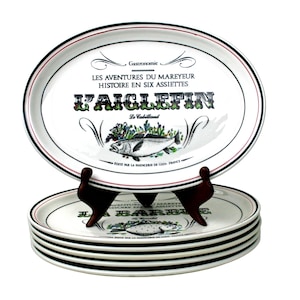 May include: A stack of white oval plates with black and red trim, featuring text and a fish illustration. The plates are displayed on a dark brown stand. The text on the plates reads "LES AVENTURES DU MAREYEUR HISTOIRE EN SIX ASSIETTES" and "L'AIGLEFIN".