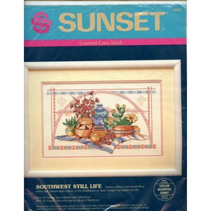 May include: A Sunset counted cross stitch kit titled "Southwest Still Life." The framed artwork depicts a still life of Southwestern pottery, cacti, and flowers. The kit includes cotton embroidery thread and a 14-count ivory Aida cloth. The frame size is 20" x 14" with mat.