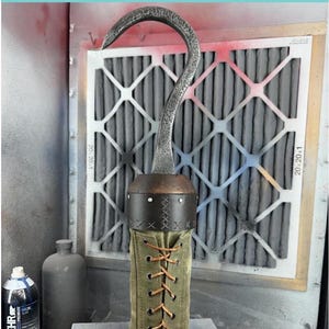 Puede incluir: Un gancho de metal con una envoltura de cuero marrón y una bota de tela verde. El gancho está unido a la bota con cordones marrones. El gancho está delante de un filtro de aire gris.