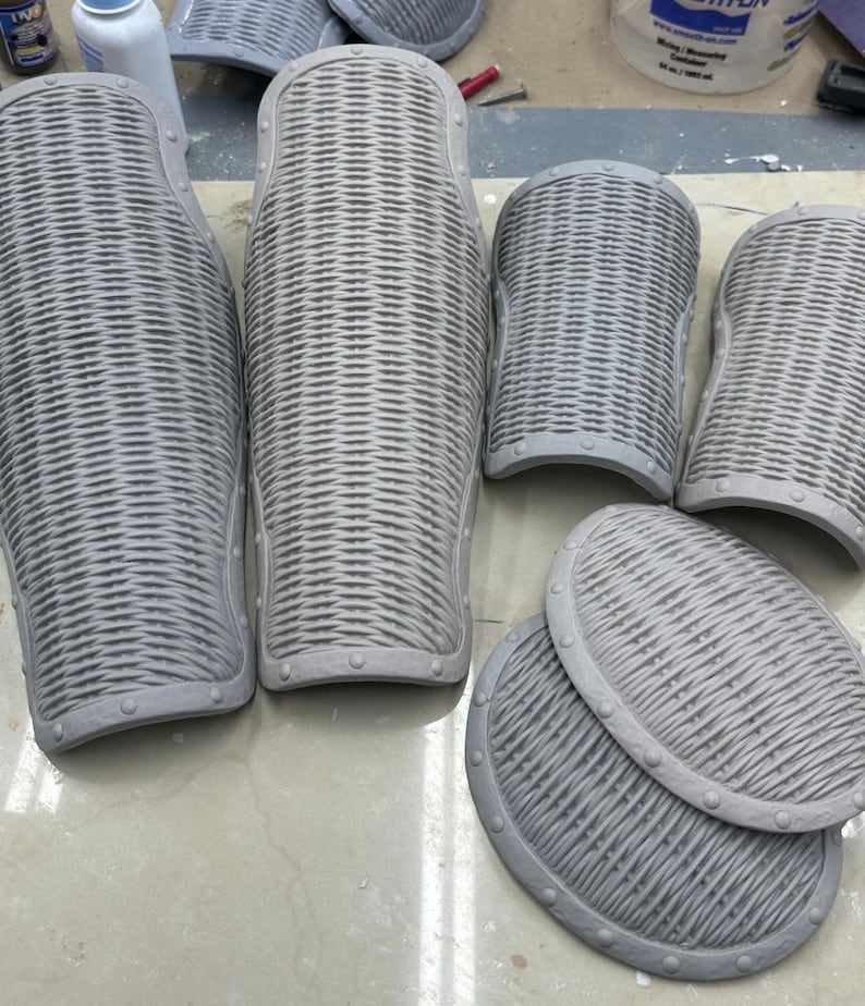 May include: Six gray, textured, foam pieces of armor. The pieces are shaped like leg guards and shields. The pieces have a woven pattern and are decorated with metal rivets.