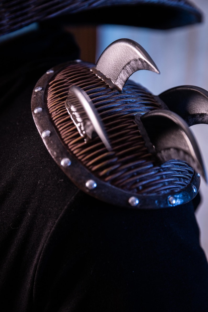 May include: Close-up of a black leather shoulder pad with a metal plate and spikes. The plate has a woven pattern and is secured with silver rivets.