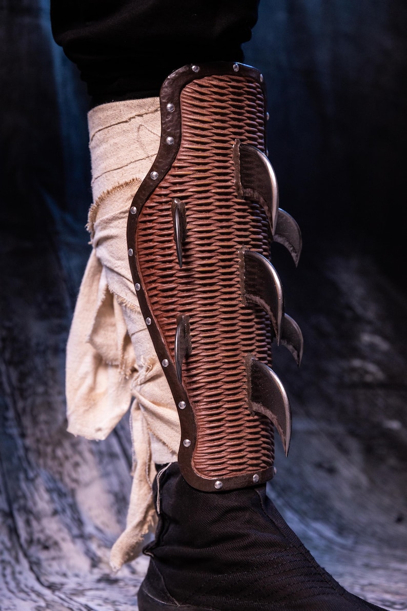 May include: A brown leather shin guard with metal spikes. The guard is worn on a leg and has a woven pattern.