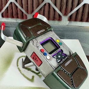 May include: A green and brown handheld device with a digital display and buttons. The device has a silver metal frame and a textured brown top. The device has a call button, a menu button, and a speaker icon.
