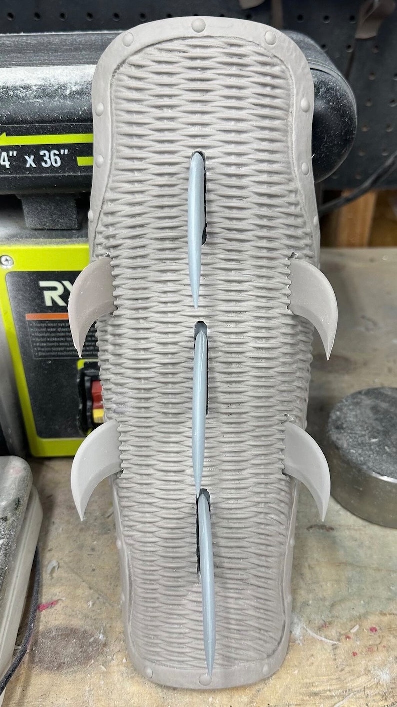 May include: A gray, textured, rectangular object with five pointed spikes protruding from the surface. The object has a raised border with small circular indentations.