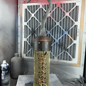 Puede incluir: Un gancho de metal con una correa de cuero marrón y una cubierta de lona verde. El gancho está asegurado con cordones marrones.