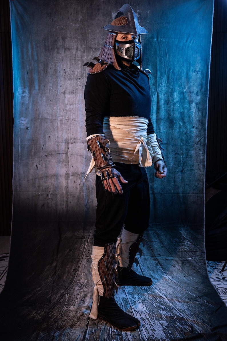 May include: A person dressed as a ninja warrior wearing a black hooded shirt, white bandages, and a silver helmet with a black mask. The ninja is wearing brown leather armor with spikes on the forearms and shins. The ninja is standing on a wooden floor in front of a blue and gray backdrop.