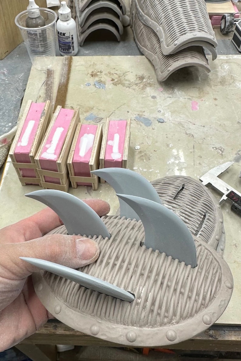 May include: A close-up of a gray, textured, round object with several gray, pointed, fin-like shapes attached to the top. The object is being held in a person's hand.