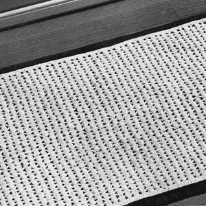 Op de afbeelding: Een close-up van een witte, gehaakte loper met een herhalend geometrisch patroon. De rechthoekige loper ligt op een donker houten oppervlak. Het haakwerk heeft een open, getextureerd ontwerp.