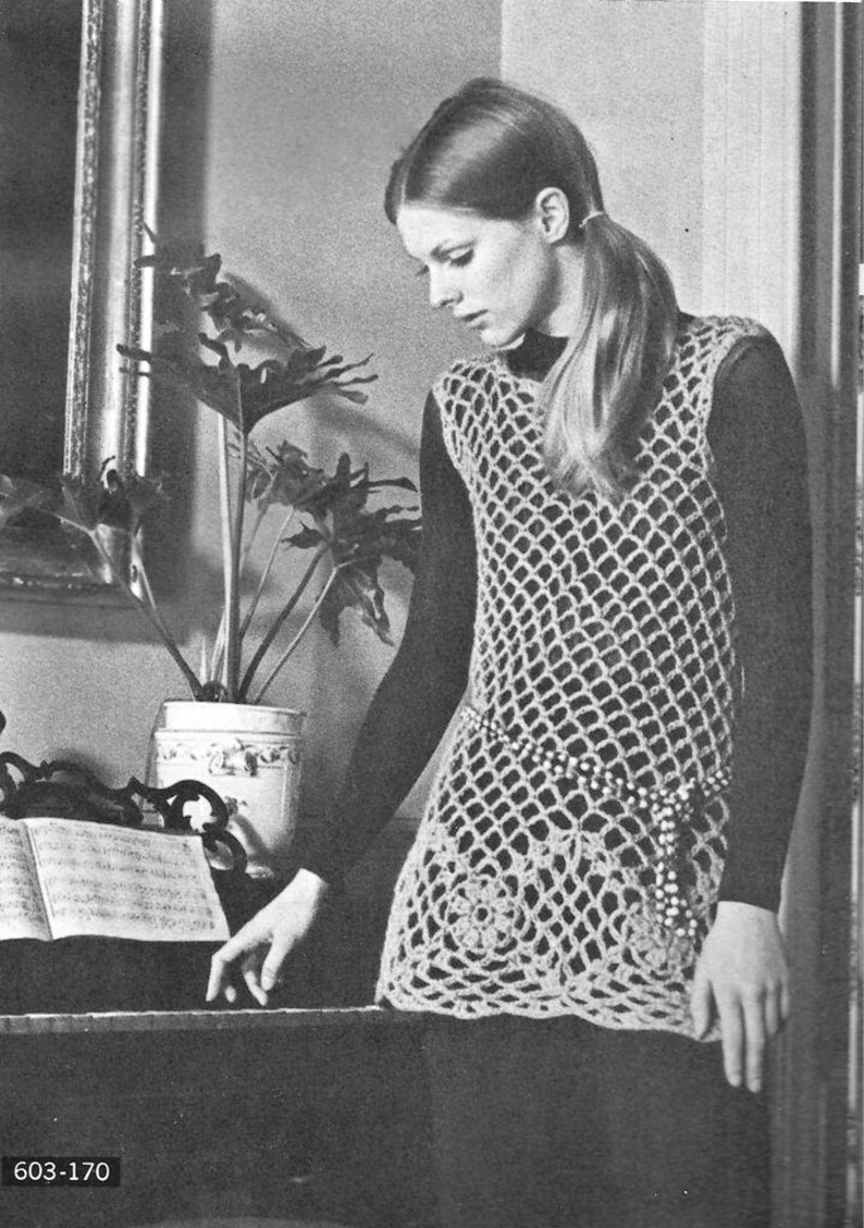 May include: A woman wearing a crocheted sleeveless top with a beaded belt. The top is made of a delicate, open-weave pattern. The woman is standing in front of a plant and a mirror.