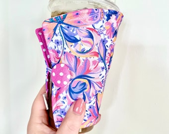 May include: A floral print coffee cup sleeve in pink, purple, and blue. The sleeve features a vibrant pattern of flowers and leaves on a white background. A pink polka dot flap secures the sleeve around the cup.