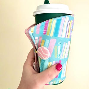 May include: A teal fabric coffee cup sleeve with a colorful book pattern. The sleeve is secured with a pink button and wraps around a green coffee cup with a white lid. The design features vertical stripes in various colors.