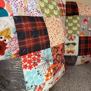 May include: Two patchwork throw pillows with a variety of fabric patterns. The patterns include floral, plaid, and butterfly designs in shades of red, orange, green, and brown. The pillows are resting on a gray couch.