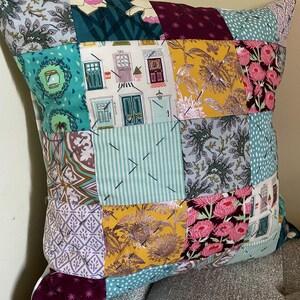 May include: A patchwork cushion with a variety of fabric patterns and colours. The cushion features squares of teal, yellow, pink, and purple fabrics with floral, door, and geometric designs. The cushion is resting on a grey upholstered surface.