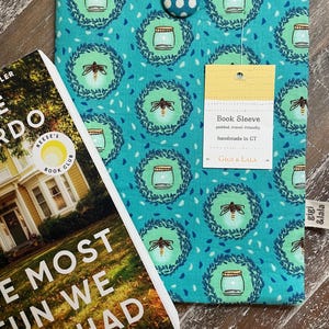 May include: A teal book sleeve with a pattern of jars and fireflies, paired with a book titled "The Most Fun We Ever Had." The sleeve has a button closure and a tag that reads "Book Sleeve." The book cover features a yellow house.