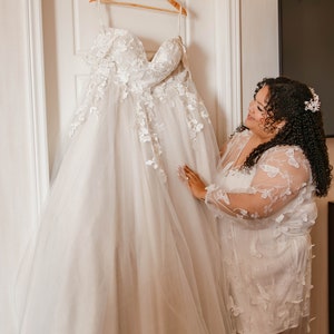 May include: An ivory wedding dress with floral lace appliqués hangs on a wooden hanger labelled "Bride". The dress is displayed on a white door. A woman in a white butterfly-patterned robe looks at the dress.