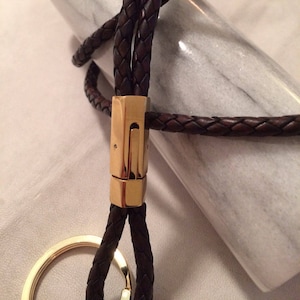 Könnte beinhalten: Ein brauner Leder-Schlüsselanhänger mit einem goldenen Metallverschluss und einem Schlüsselring. Das Leder ist geflochten und hat eine dunkelbraune Farbe.